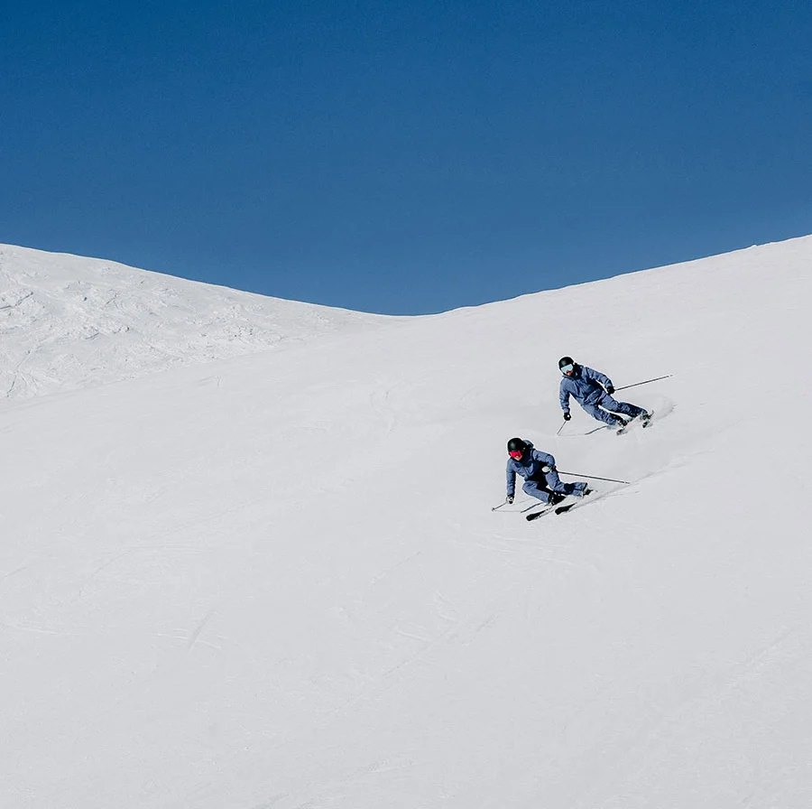 Zwei Skifahrer auf einem weiten Schneehang unter blauem Himmel